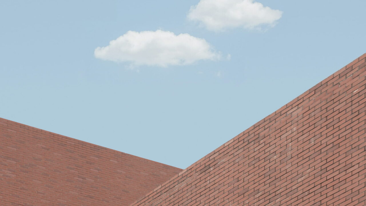 The top edge of a brick building against a pale blue sky with two small clouds.