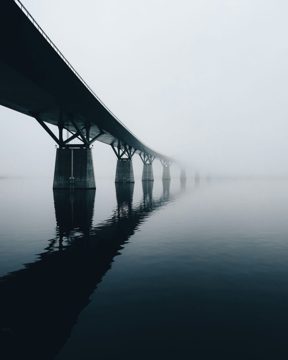 A long concrete bridge on pillars disappearing into fog over still water.