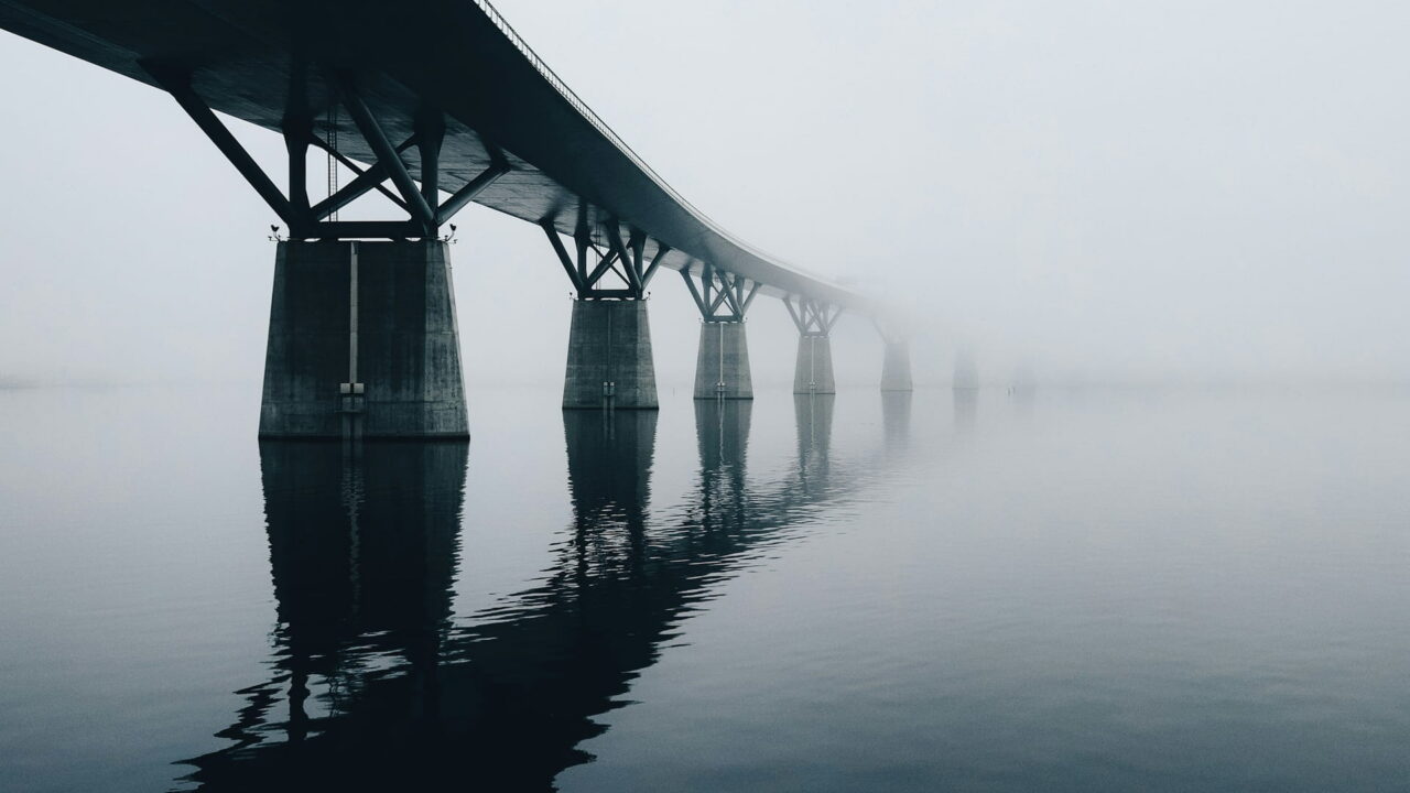 A long concrete bridge on pillars disappearing into fog over still water.