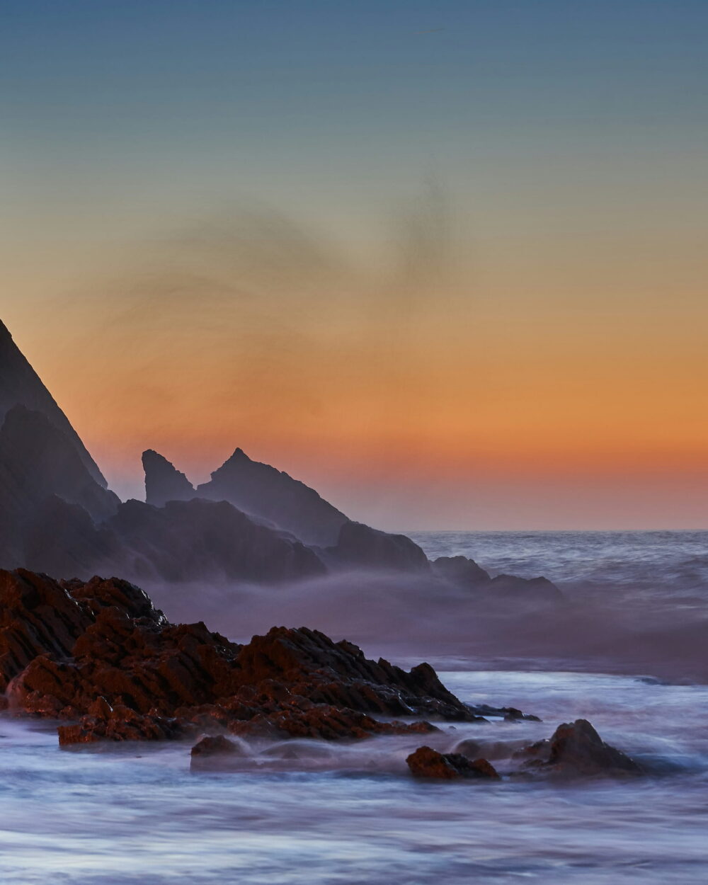 Dark sea stacks and a cliff in mist against an orange dusk sky.