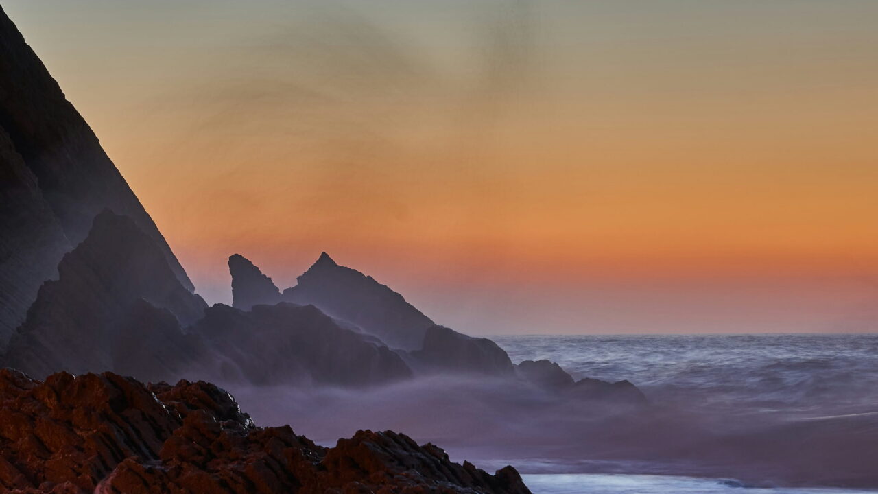 Dark sea stacks and a cliff in mist against an orange dusk sky.