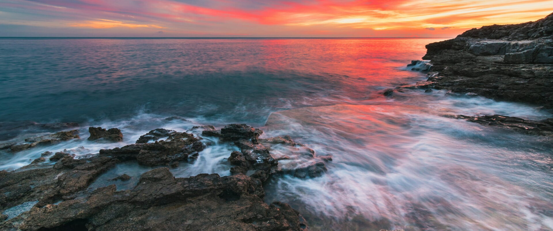 Waves washing over flat rocks under a red and orange sunset sky.