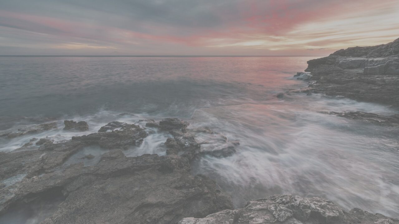 The same sunset over waves and flat rocks, flat and pale, as the camera recorded it before printing.