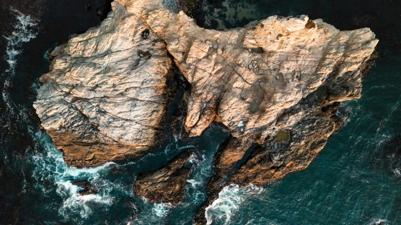 An aerial view of a pale rock island in dark green sea, ringed with white foam.
