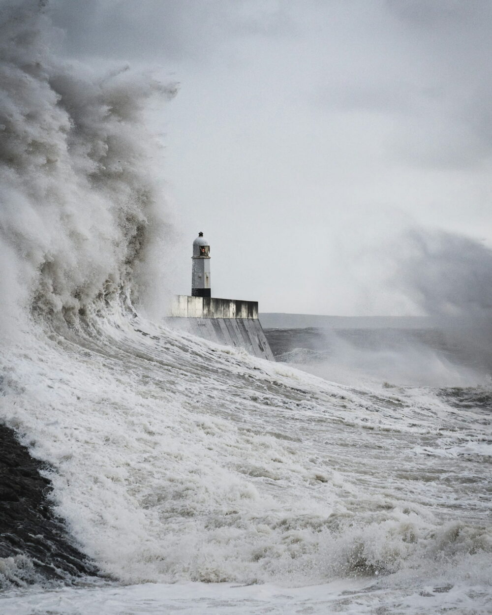 A huge wave breaking around a lighthouse at the end of a harbor wall.