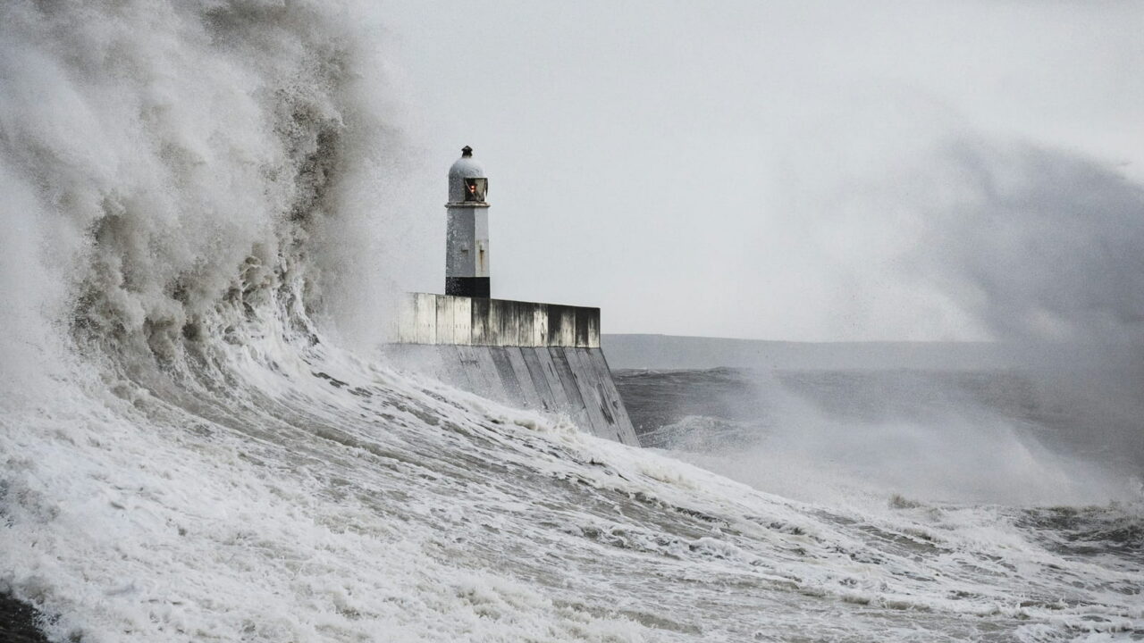 A huge wave breaking around a lighthouse at the end of a harbor wall.