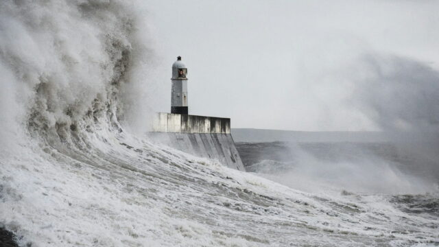 A huge wave breaking around a lighthouse at the end of a harbor wall.