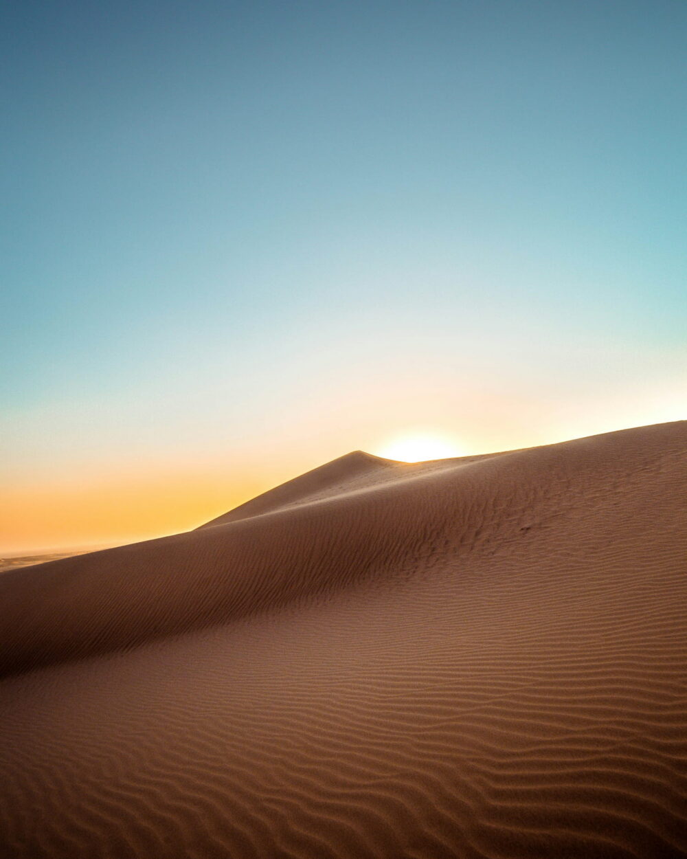 The sun rising behind the sharp edge of a sand dune under a clear sky.