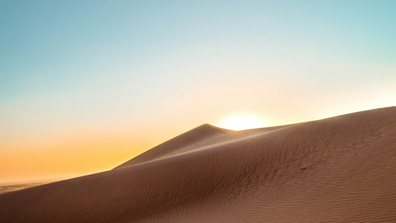 The sun rising behind the sharp edge of a sand dune under a clear sky.
