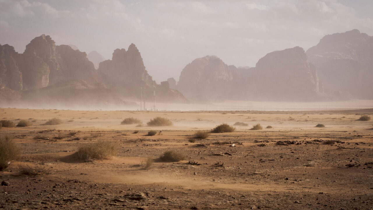 Dust blowing across a dry valley floor in front of pale rock towers.