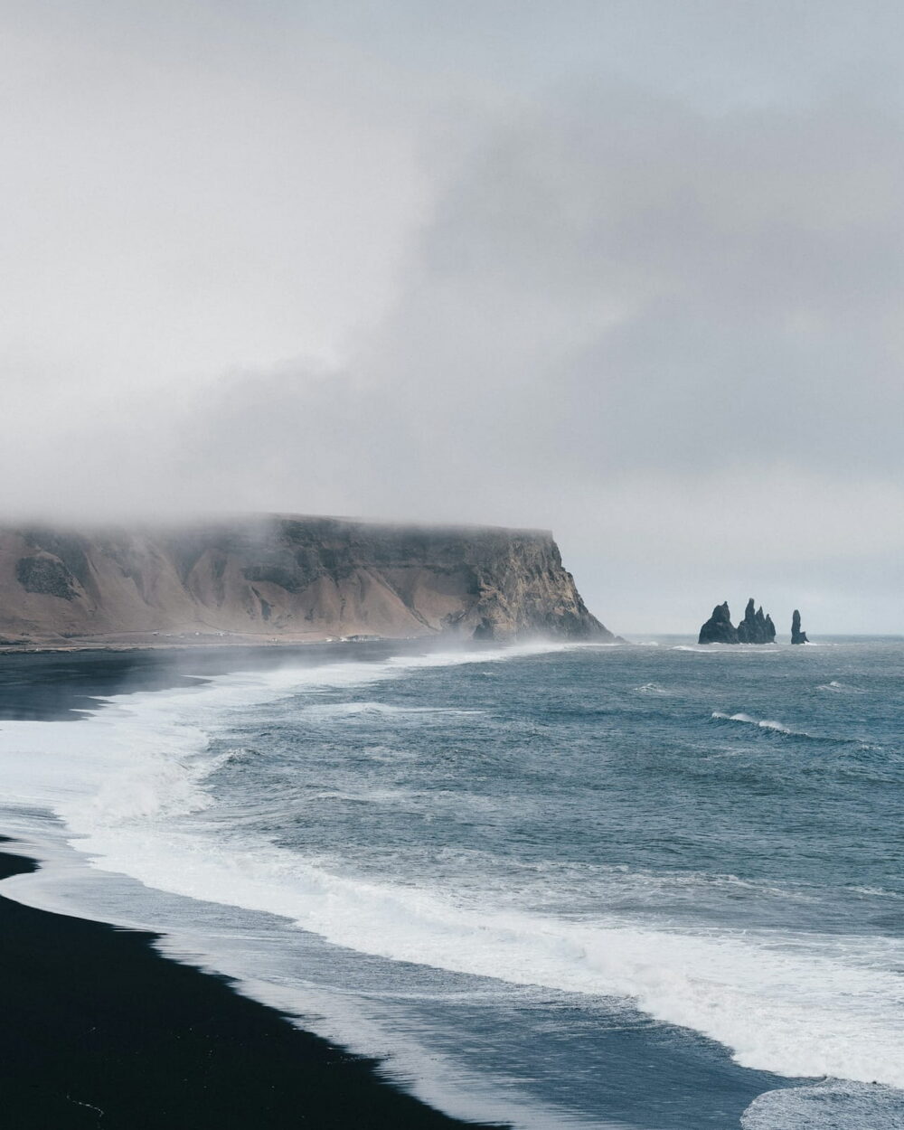 Waves running up a black sand beach below misty sea cliffs and two rock stacks.