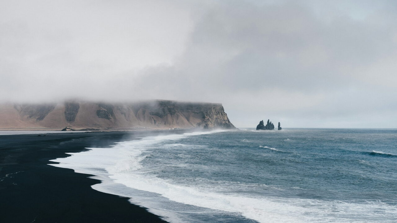 Waves running up a black sand beach below misty sea cliffs and two rock stacks.
