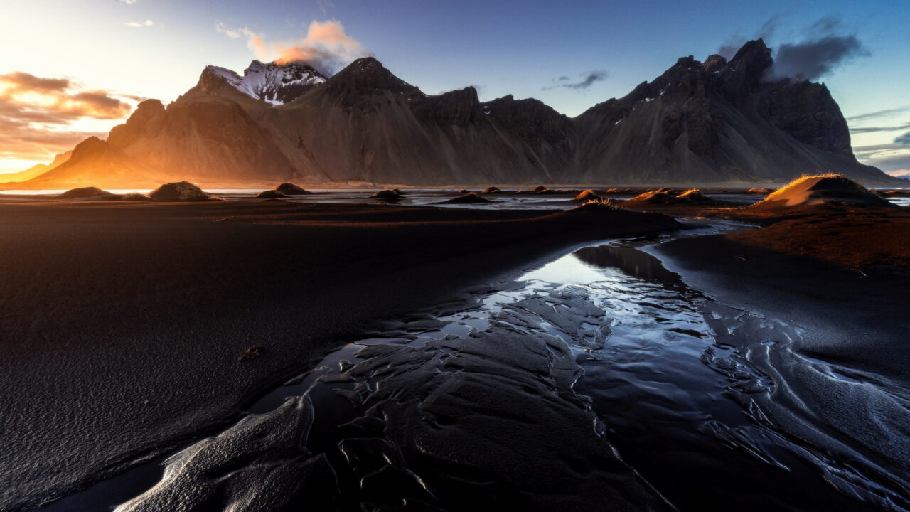 Low sun breaking over a sharp mountain ridge, with water shining in channels across black dunes.