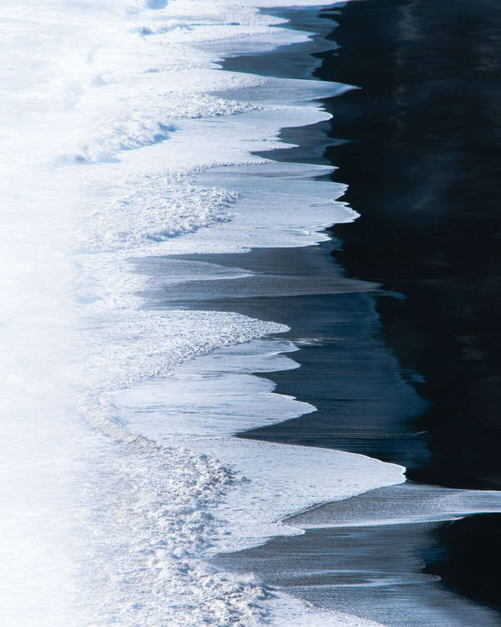 Seen from above, white surf meets a band of black sand in a clean diagonal line.