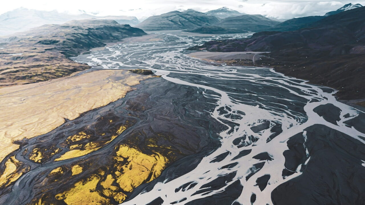 An aerial view of a pale braided river spreading across dark gravel plains toward the mountains.