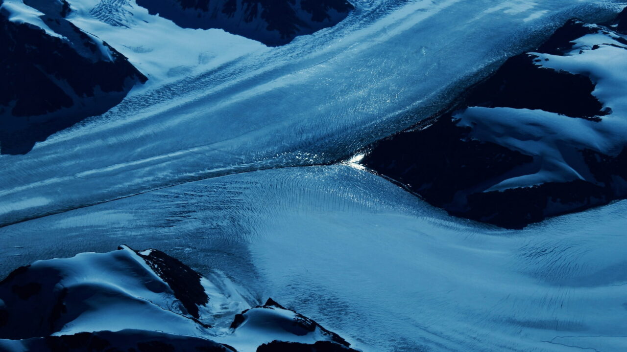 An aerial view of a blue glacier tongue flowing between dark, snowy ridges.