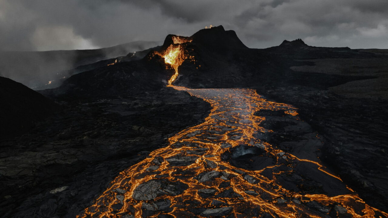 Orange lava running in glowing channels across a dark volcanic field under smoke.