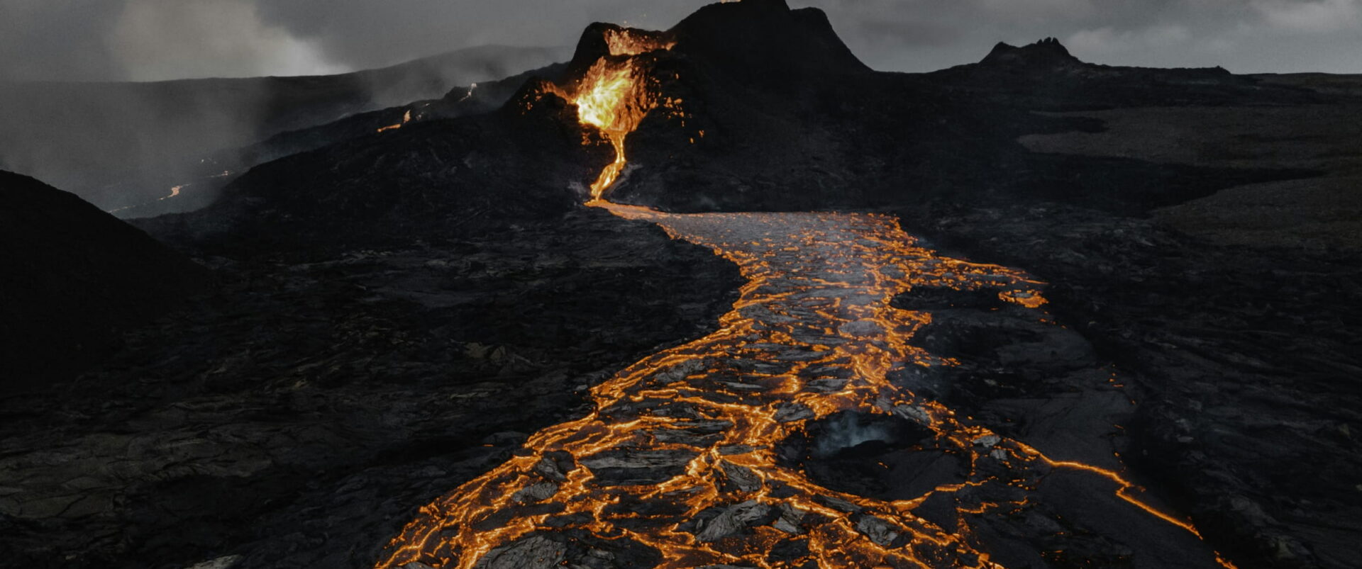 Orange lava running in glowing channels across a dark volcanic field under smoke.
