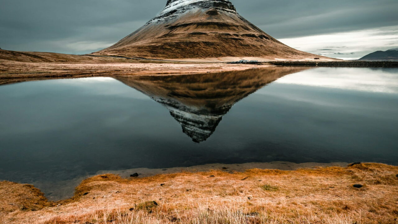 A cone-shaped, striped mountain reflected in still water above a bank of golden grass.