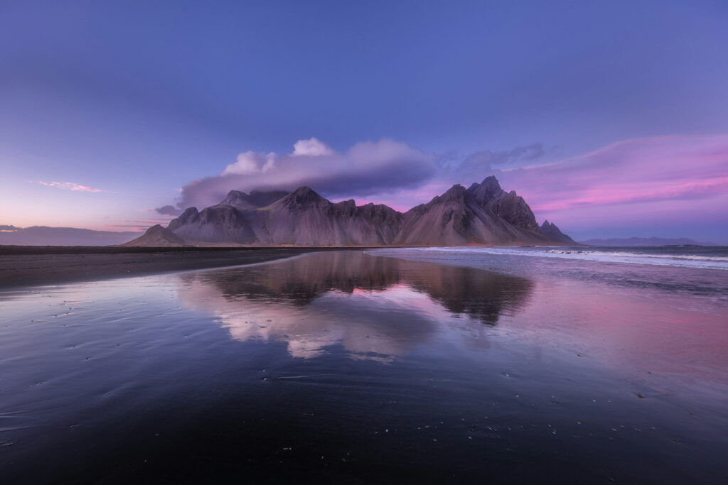 A jagged mountain range reflected in wet black sand under a pink and violet dusk sky.