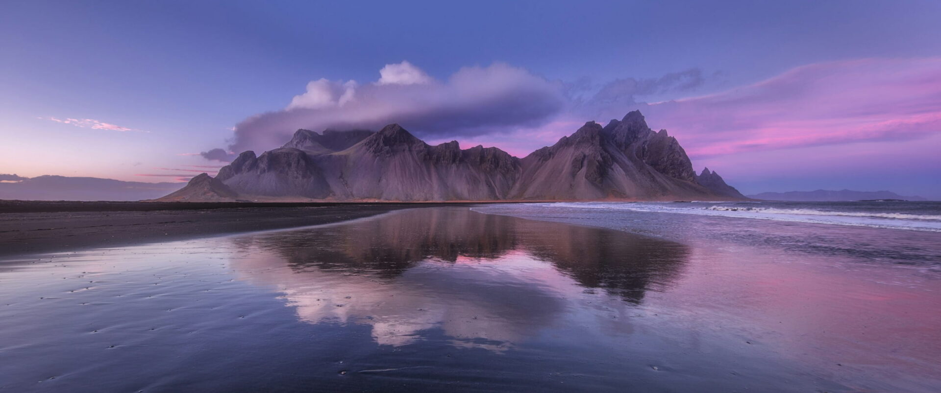 A jagged mountain range reflected in wet black sand under a pink and violet dusk sky.