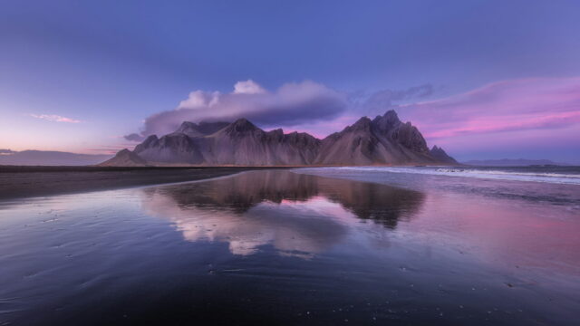 A jagged mountain range reflected in wet black sand under a pink and violet dusk sky.