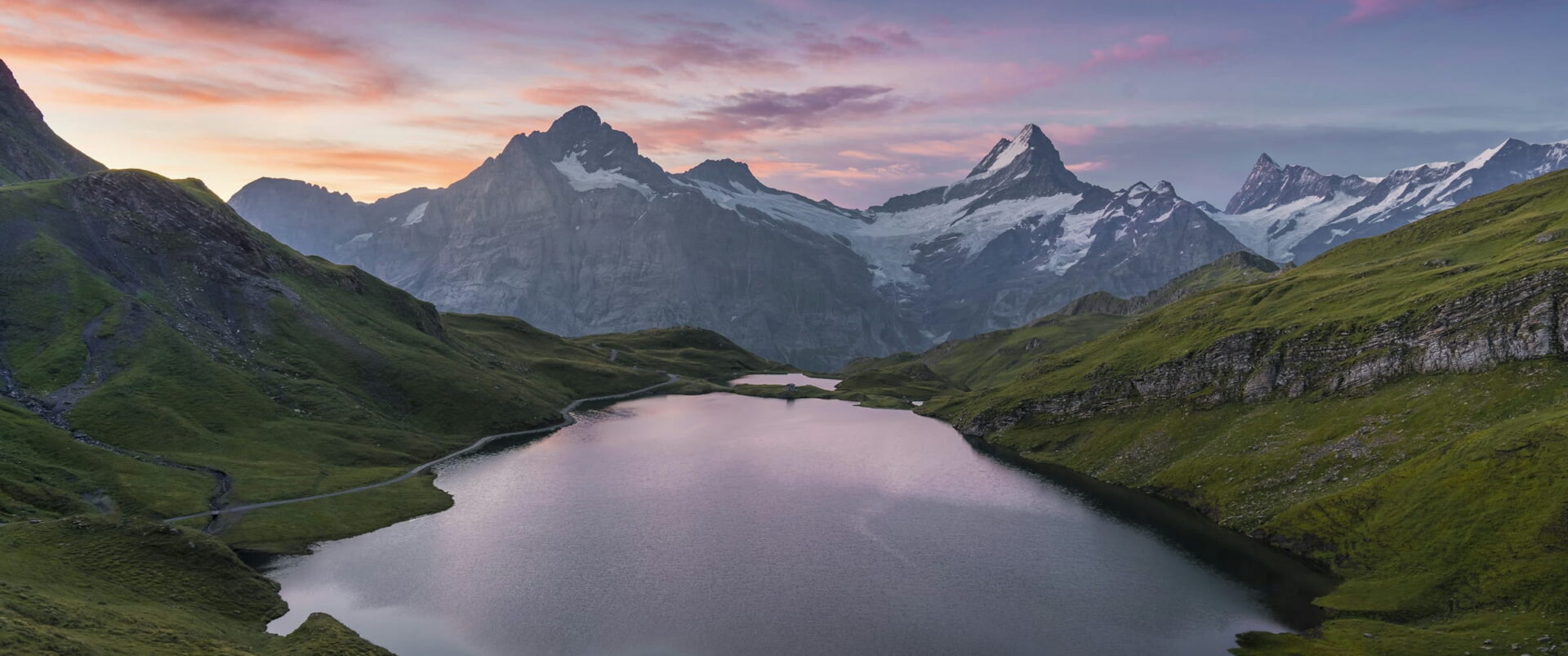 A still alpine lake in a green valley, with snowy peaks under a pink and violet dawn sky.