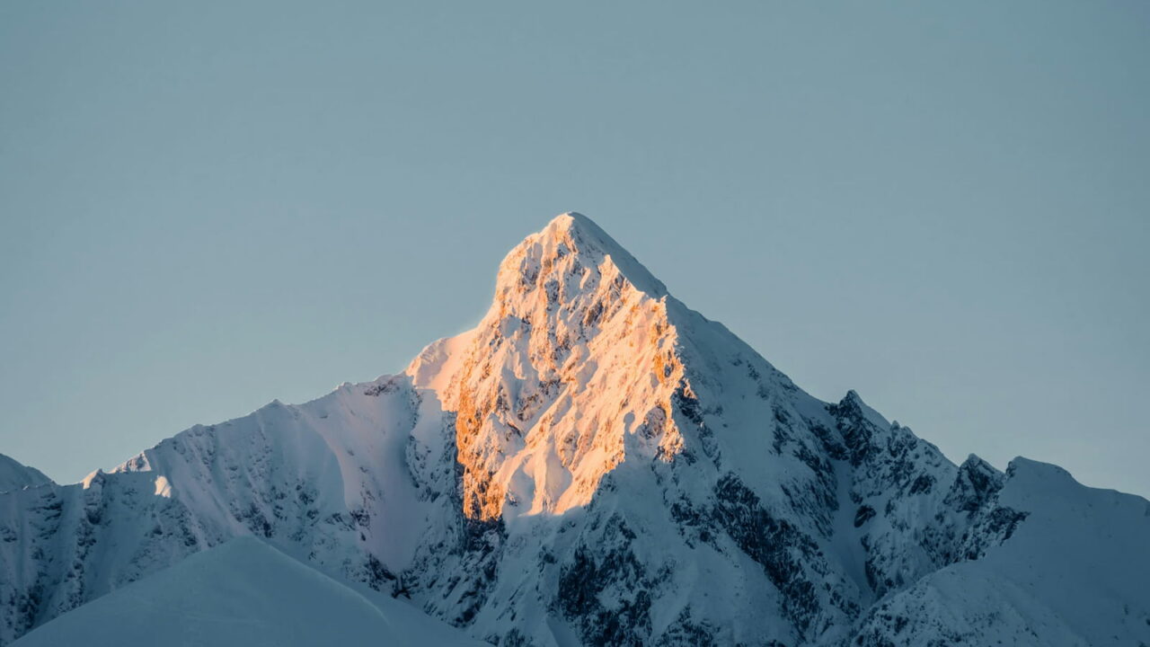 The tip of a snow peak catching orange first light above a blue, shadowed slope.