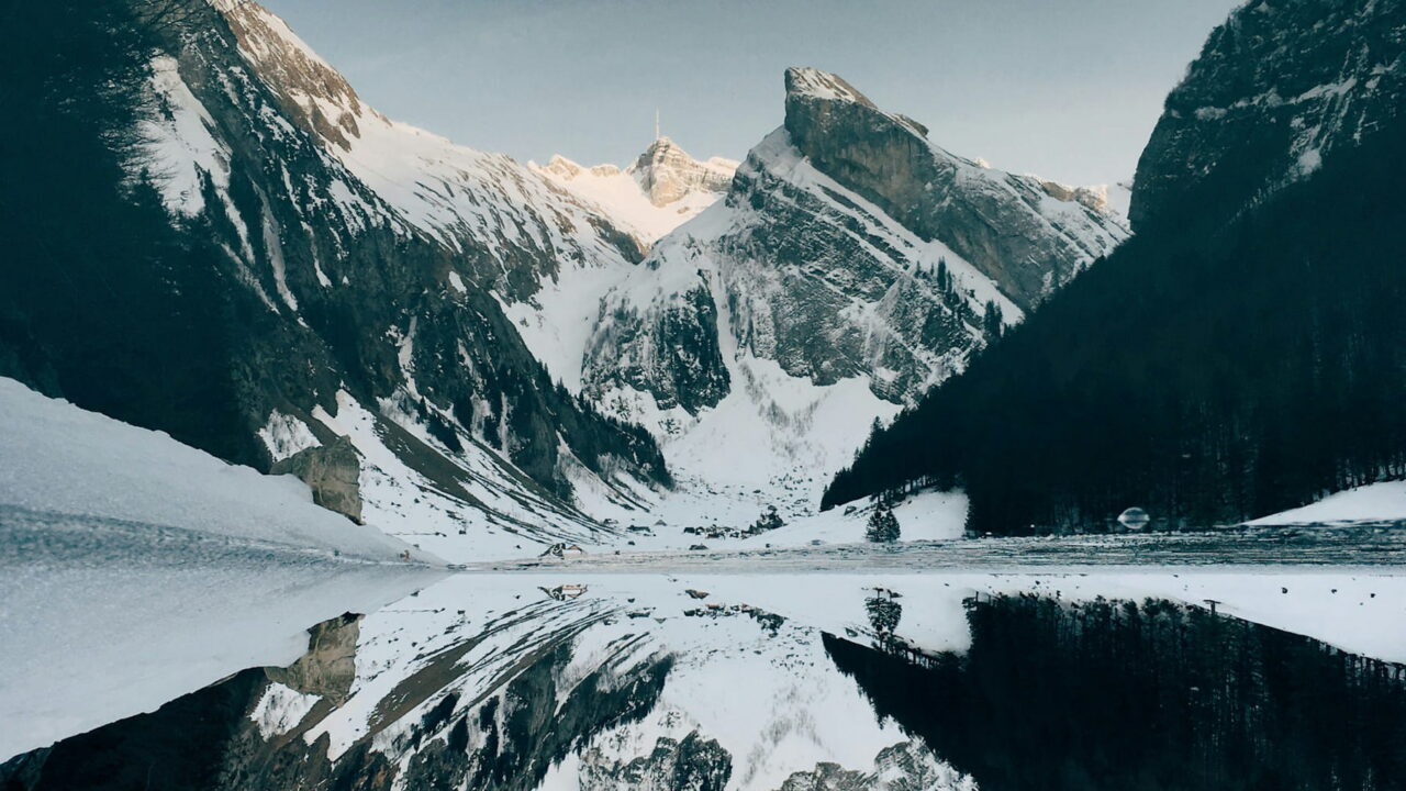 Jagged, snowy mountains reflected in a still lake between dark cliffs.
