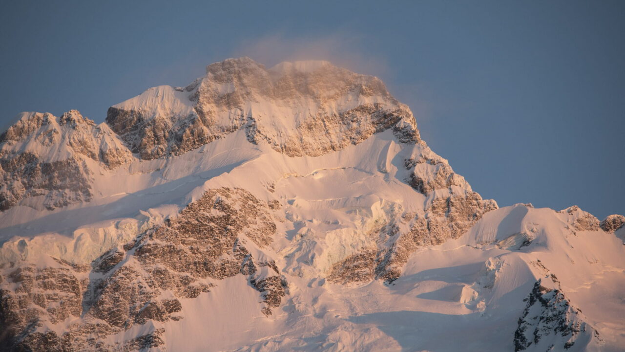 A massive snowy rock face lit warm by the evening sun, with snow blowing off the top.