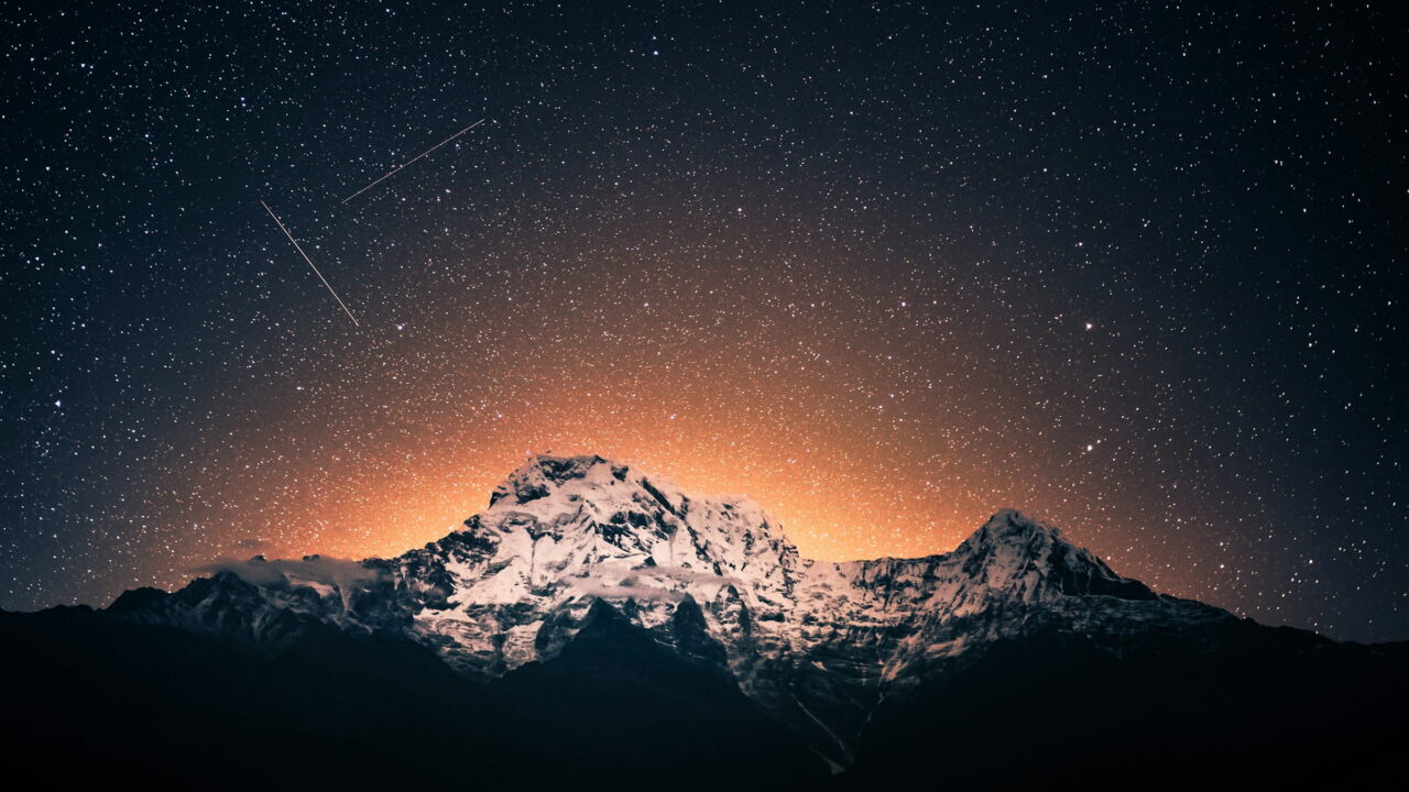 A snow peak lit by a warm glow on the horizon, under a sky full of stars and one meteor.