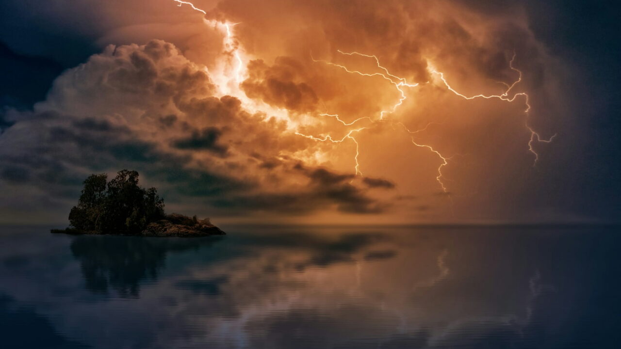 Lightning running through orange storm clouds above a small wooded island and its reflection.