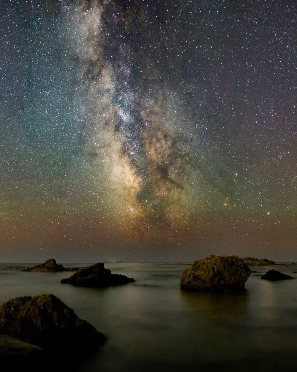 The Milky Way over a calm sea with dark boulders and a faint green glow on the horizon.