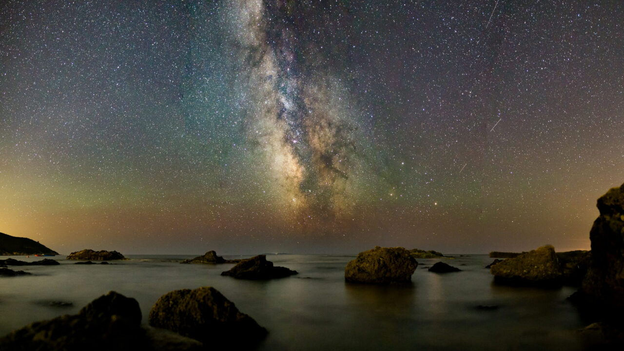 The Milky Way over a calm sea with dark boulders and a faint green glow on the horizon.