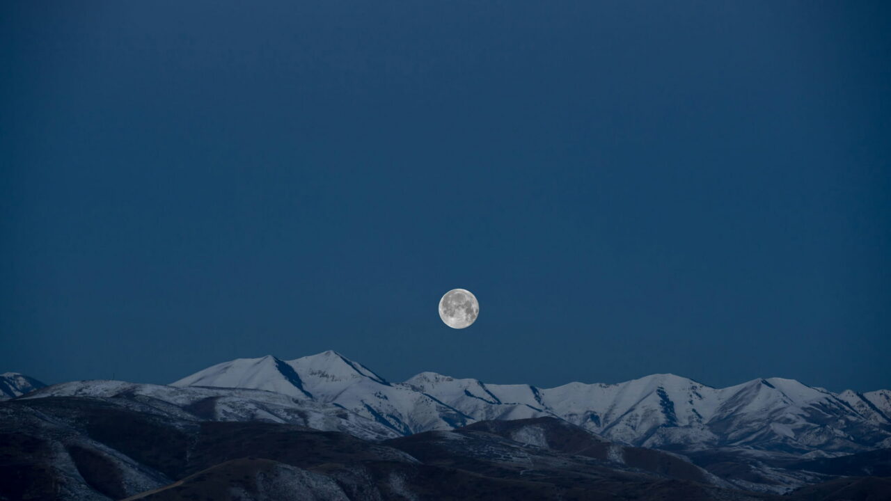 A small full moon over a snow-capped mountain range in a deep blue sky.