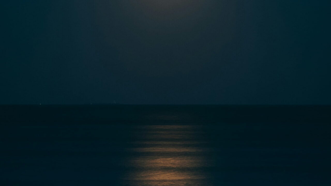 An orange full moon laying a path of light across a dark, flat sea.